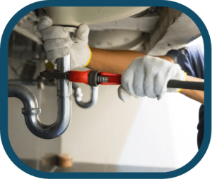 A person wearing white gloves uses a red pipe wrench to tighten or loosen a metal pipe under a sink during a Kitchen Plumbing Repair in Denver. | High 5 Plumbing, Heating & Cooling