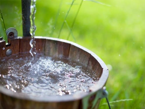 A wooden bucket is being filled with clear water from a small stream, with splashes visible. The background is grassy and out of focus, suggesting an outdoor setting. | High 5 Plumbing, Heating & Cooling