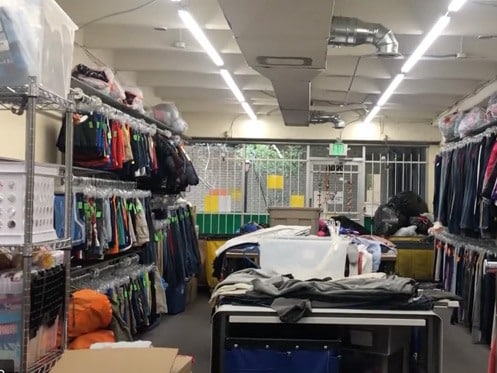 A room with fluorescent lighting containing clothing racks filled with various garments, tables with folded clothes, metal shelves with storage bins, and barred windows at the back. | High 5 Plumbing, Heating & Cooling