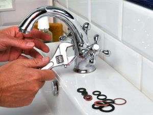 A person uses a wrench to repair a chrome faucet on a white sink, with various rubber washers and gaskets laid out on the countertop nearby. | High 5 Plumbing, Heating & Cooling