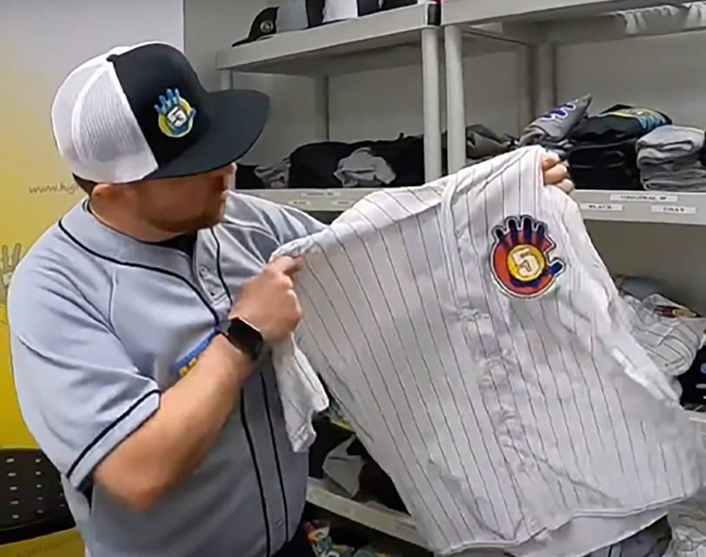 A man wearing a baseball jersey and cap holds up a white pinstriped baseball shirt with a colorful "5" logo patch, standing in front of shelves with folded clothes. | High 5 Plumbing, Heating & Cooling