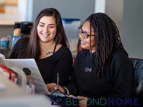 Two women sit side by side at a desk, looking at documents and smiling. One has long straight hair, the other has long dreadlocks and glasses. The BeyondHome logo appears at the bottom of the image. | High 5 Plumbing, Heating & Cooling