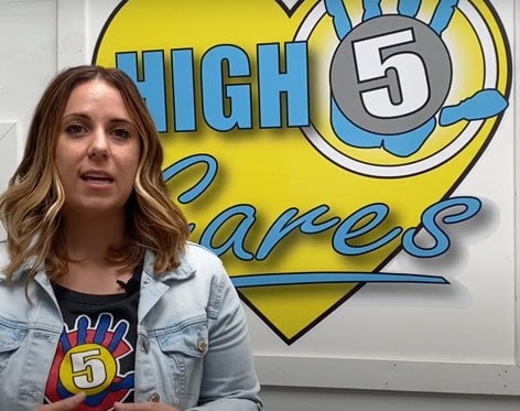 A woman with shoulder-length hair stands in front of a sign reading "High 5 Cares" with a heart and hand graphic. She wears a denim jacket and a shirt with the same "High 5" logo. | High 5 Plumbing, Heating & Cooling