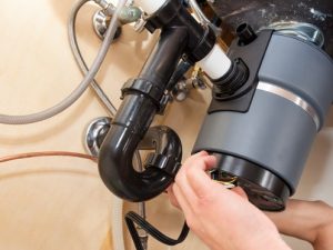 A person installs or repairs a garbage disposal under a kitchen sink, connecting wires to the unit and surrounded by pipes and plumbing fixtures. | High 5 Plumbing, Heating & Cooling