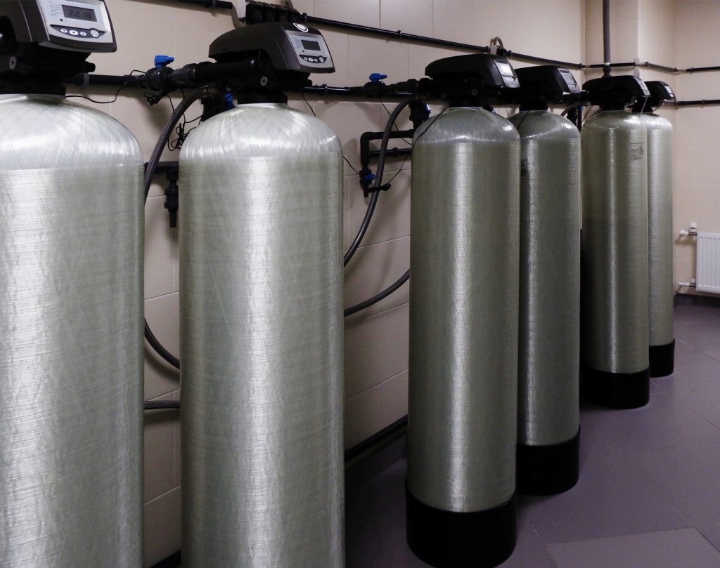 Five large industrial water filtration tanks with digital control panels are lined up against a wall, interconnected by pipes and hoses in a clean, tiled utility room. | High 5 Plumbing, Heating & Cooling