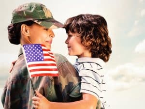 A woman in military uniform holds a young boy in her arms. The boy holds a small American flag, and they look at each other, both smiling. The background is mostly sky with clouds. | High 5 Plumbing, Heating & Cooling