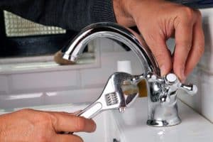 A person uses a wrench to tighten or adjust a metal faucet handle on a bathroom sink with white tile in the background, following faucet tips to avoid leaks and prevent a leaking faucet. | High 5 Plumbing, Heating & Cooling