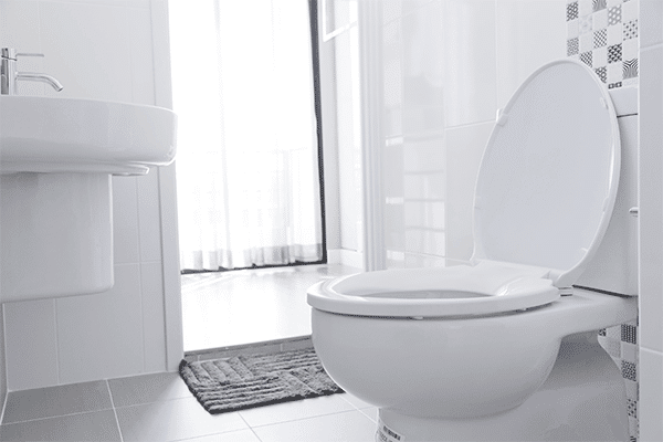 A white toilet with the lid open sits beside a white sink in a clean, modern bathroom. Sunlight streams through a partially open door with sheer curtains. Eco-friendly bathroom products complement the space, with a grey floor mat in front of the toilet. | High 5 Plumbing, Heating & Cooling