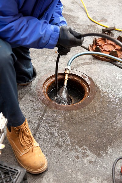 A person wearing a blue jacket, black gloves, and tan work boots is performing sewer repair, holding a hose and inserting it into an open drain or manhole on a concrete surface. Water and equipment are visible around the opening. | High 5 Plumbing, Heating & Cooling