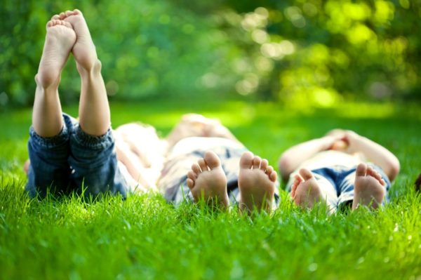 Three children lie barefoot on green grass, their feet in the foreground near an outdoor faucet. The blurred greenery and sunlight in the background evoke a relaxed outdoor setting. The children are wearing jeans. | High 5 Plumbing, Heating & Cooling