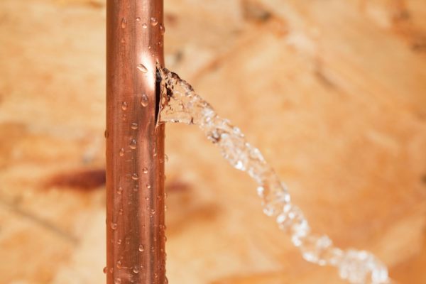 Water streams out of a crack in a vertical copper pipe against a blurred tan background, highlighting the importance of winter plumbing tips to prevent frozen pipes and avoid pipes bursting during cold weather. | High 5 Plumbing, Heating & Cooling