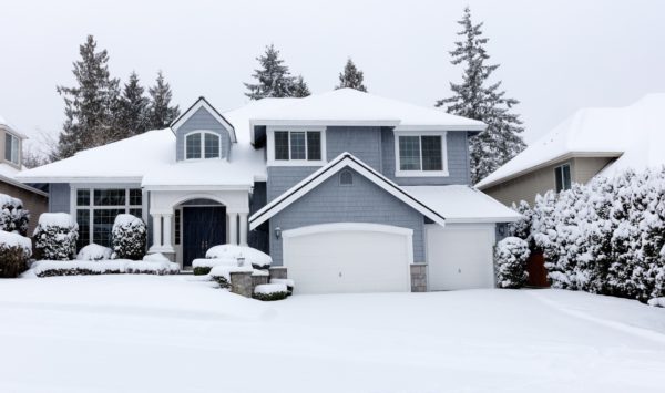 A two-story suburban house with snow covering the roof, driveway, and yard. Evergreen trees and bushes are blanketed in snow, while the overcast sky hints at a recent snowfall—perfect weather to check and winterize plumbing systems. | High 5 Plumbing, Heating & Cooling