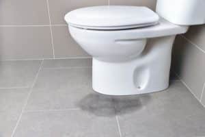 A white toilet sits against a tiled wall on a gray tiled floor. A visible wet stain near the base suggests potential toilet maintenance is needed to prevent further issues. | High 5 Plumbing, Heating & Cooling