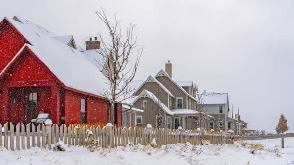 Several houses, including a prominent red one, are lined up behind a white picket fence on a snowy day. Snow blankets the roofs and ground—an ideal reminder to take steps for pipe freezing prevention this winter. | High 5 Plumbing, Heating & Cooling