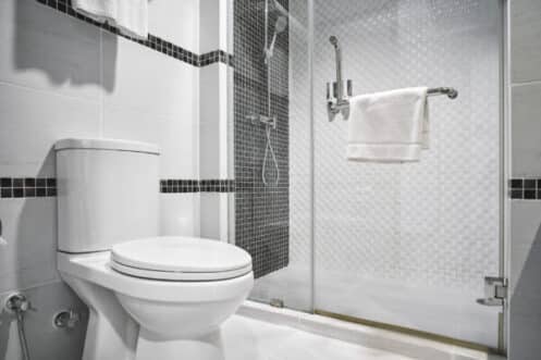 A modern bathroom with a white toilet, glass-enclosed shower, wall-mounted towel rack with a white towel, and stylish black-and-white tile accents showcases how indoor plumbing has evolved since the first people began to invent sanitation systems. | High 5 Plumbing, Heating & Cooling