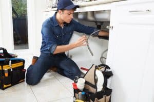 A plumber wearing a blue shirt, jeans, and cap is kneeling on a kitchen floor, working under a sink with plumbing essentials and supplies in tool bags nearby. | High 5 Plumbing, Heating & Cooling