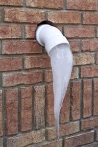 A large icicle hangs from the end of a white PVC pipe sticking out of a brick wall, formed where drains release water after heavy rainfall. | High 5 Plumbing, Heating & Cooling