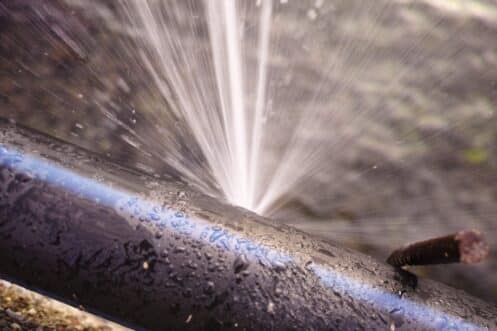 A close-up of a black pipe with a blue stripe, spraying water forcefully from a pipes burst. Water droplets are visible on the pipe, and a rusty metal rod is nearby. | High 5 Plumbing, Heating & Cooling