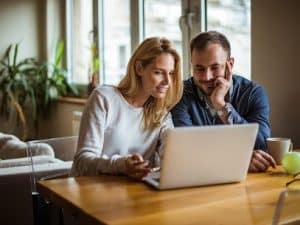 A woman and a man sit at a wooden table, looking at a laptop screen together. The man holds a mug and an apple is on the table as they research the benefits of installing a yard hydrant. Large windows and a plant are in the background. | High 5 Plumbing, Heating & Cooling