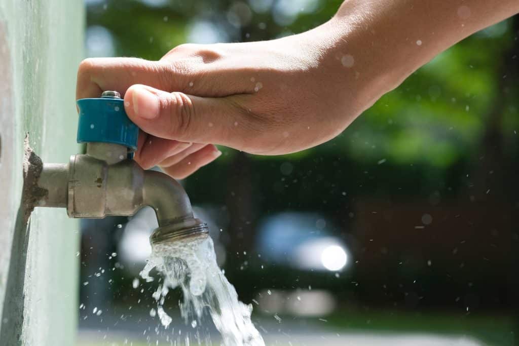 A person’s hand is turning on a blue-handled outdoor water faucet, with water flowing out of the tap and droplets splashing—highlighting reliable access, much like gas plumbing during a power outage. The background is blurred greenery and light. | High 5 Plumbing, Heating & Cooling