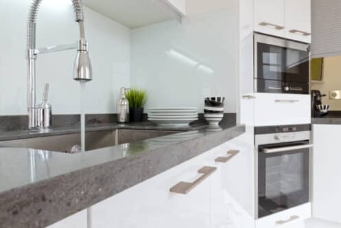 Modern kitchen with white cabinets, a built-in oven and microwave, a stainless steel sink with a high-arc faucet showing hard water signs, and stacked dishes and cups on the countertop near a green potted plant. | High 5 Plumbing, Heating & Cooling