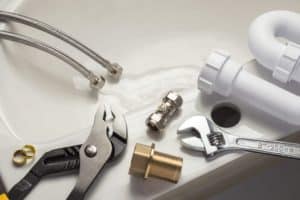 Plumbing tools and materials for bathtub repair are laid out on a white surface next to a sink. Items include a wrench, pliers, pipe fittings, valves, braided hoses, a plastic pipe, and brass components—everything needed to fix bathtub cracks. | High 5 Plumbing, Heating & Cooling