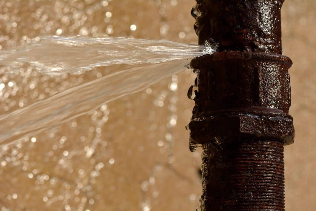 Water sprays from a crack in a rusty metal pipe, signaling a pipe burst. The blurred, water-stained background highlights the need for emergency plumbing. | High 5 Plumbing, Heating & Cooling