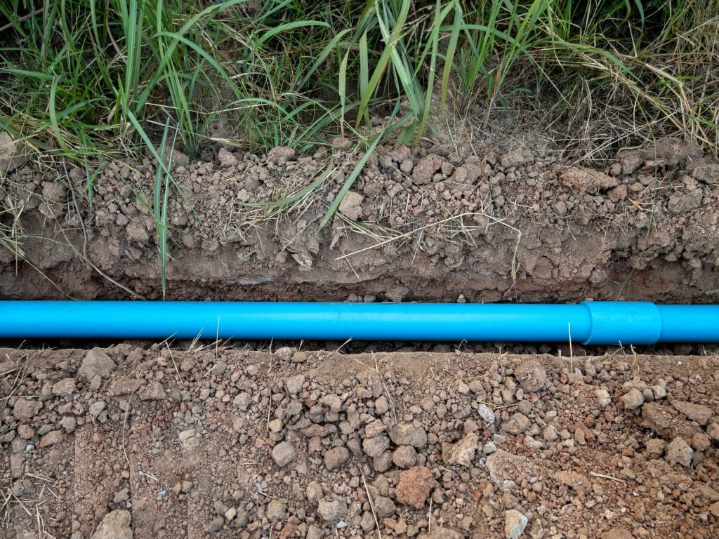 A blue plastic pipe, potentially vulnerable to sewer line deterioration, is laid horizontally in a narrow trench filled with loose soil and surrounded by grass and dirt. | High 5 Plumbing, Heating & Cooling