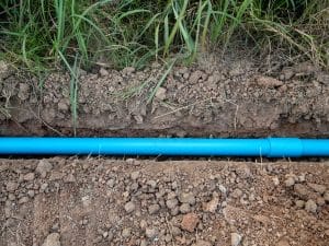A blue plastic pipe, potentially vulnerable to sewer line deterioration, is laid horizontally in a narrow trench filled with loose soil and surrounded by grass and dirt. | High 5 Plumbing, Heating & Cooling
