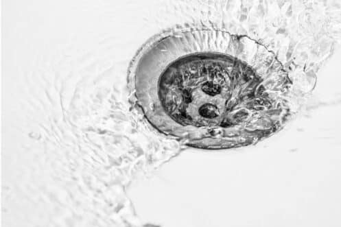Close-up of water swirling around and draining into a metal sink drain, with ripples and reflections visible on the white sink surface near a subtle sink crack. | High 5 Plumbing, Heating & Cooling