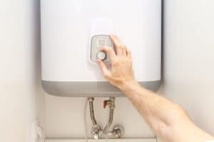 A person’s hand is adjusting the temperature dial on a white wall-mounted water heater, helping to extend its life expectancy, with visible water pipes and fittings underneath the unit. | High 5 Plumbing, Heating & Cooling