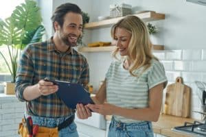A man in a plaid shirt with a tool belt and a woman in a striped shirt stand in a kitchen, both smiling and looking at a clipboard as they discuss the latest plumbing trends for 2024. | High 5 Plumbing, Heating & Cooling