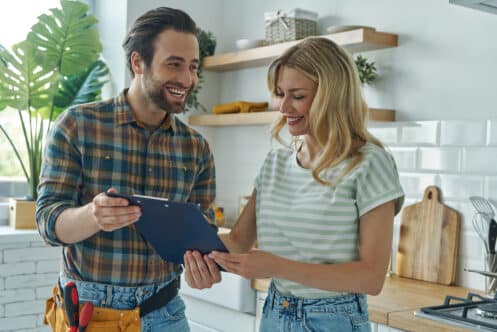 A man in a plaid shirt with a tool belt and a woman in a striped shirt stand in a kitchen, both smiling and looking at a clipboard as they discuss the latest plumbing trends for 2024. | High 5 Plumbing, Heating & Cooling