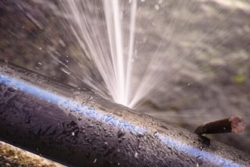 A close-up of a black pipe with a blue stripe spraying water forcefully from a leak, illustrating why pipes burst. The background is blurred, and a rusty screw or nail is visible near the right side of the pipe. | High 5 Plumbing, Heating & Cooling