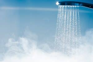A close-up of a showerhead spraying water, with visible steam rising below, indicating hot water in use. The soft blue background adds to the clean, fresh atmosphere—perfect inspiration for luxurious bathroom or shower upgrades. | High 5 Plumbing, Heating & Cooling