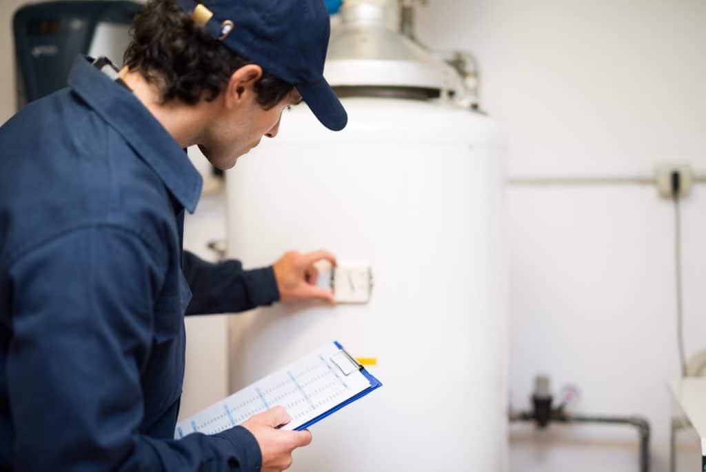 A person in a blue uniform and cap inspects a white water heater, adjusting a control or thermostat while holding a clipboard with a checklist in a utility room, ensuring all plumbing and gas line connections are safe and up to code. | High 5 Plumbing, Heating & Cooling