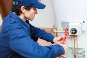 A person wearing a blue uniform and cap uses a red pipe wrench to adjust a component on a white water heater, showcasing emergency plumbing skills. Various pipes and a control panel are visible on the heater. | High 5 Plumbing, Heating & Cooling
