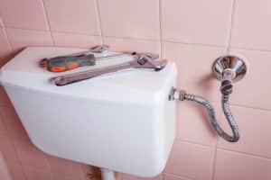 A toilet tank against a pink tiled wall with a wrench and screwdriver on top, next to a metal braided water supply line connected to a shut-off valve—different plungers like a sink plunger or toilet plunger aren't pictured but are helpful for maintenance. | High 5 Plumbing, Heating & Cooling