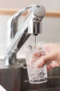 A hand holds a clear glass under a modern kitchen faucet, filling it with water filtered by a whole-home water filtration system. The background shows part of a stainless steel sink and countertop. | High 5 Plumbing, Heating & Cooling