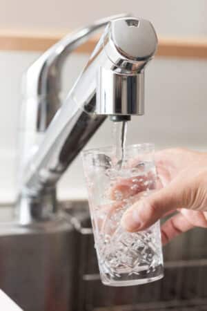 A hand holds a clear glass under a modern kitchen faucet, filling it with water filtered by a whole-home water filtration system. The background shows part of a stainless steel sink and countertop. | High 5 Plumbing, Heating & Cooling