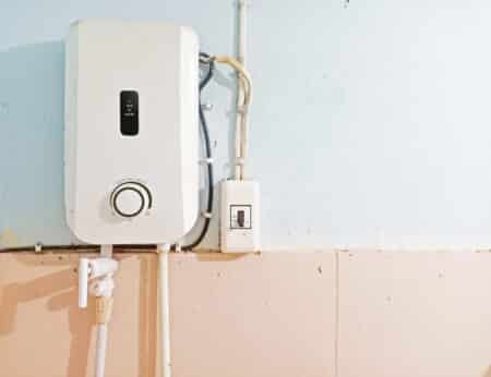 A white wall-mounted tankless water heater with visible pipes and a control panel is installed on a pale blue and beige tiled wall. An electrical switch is positioned next to the heater, highlighting easy access for maintenance. | High 5 Plumbing, Heating & Cooling