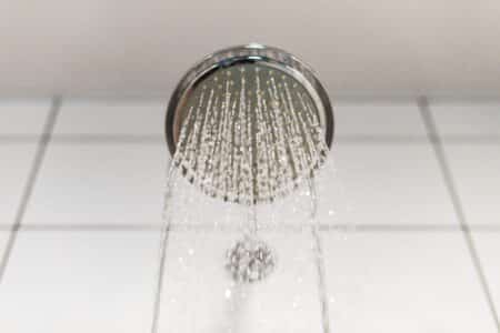 A close-up view of a round showerhead with soft water spraying down, creating a gentle flow that's kind to your hair and skin, against a background of white tiled wall. | High 5 Plumbing, Heating & Cooling