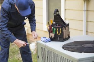 A person in a navy uniform and cap uses a screwdriver for air conditioner troubleshooting on an outdoor unit. A tool bag with various tools and a blue cloth sits on top of the unit next to them. | High 5 Plumbing, Heating & Cooling