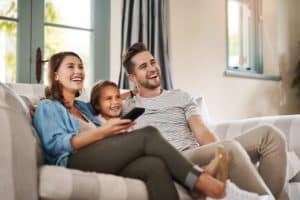 A woman, a man, and a young girl sit together on a couch in a living room, smiling and looking at something off-camera. The woman holds a remote control in their smart home, filled with natural light from energy-efficient windows behind them. | High 5 Plumbing, Heating & Cooling