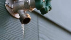 A close-up of an outdoor metal water faucet with a green handle, attached to a wall, with a small icicle hanging from the spout—an early sign of winter and a reminder of the risk of frozen pipes. | High 5 Plumbing, Heating & Cooling