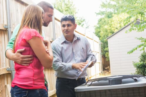 A man in a button-up shirt holds a clipboard and explains heating and cooling tips to a couple standing outside next to an outdoor AC unit near a wooden fence and shed. | High 5 Plumbing, Heating & Cooling