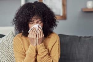 A person with curly hair wearing a yellow sweater sits on a sofa, holding a tissue to their nose, possibly reacting to poor winter indoor air quality. The background features a patterned pillow and a softly blurred interior. | High 5 Plumbing, Heating & Cooling