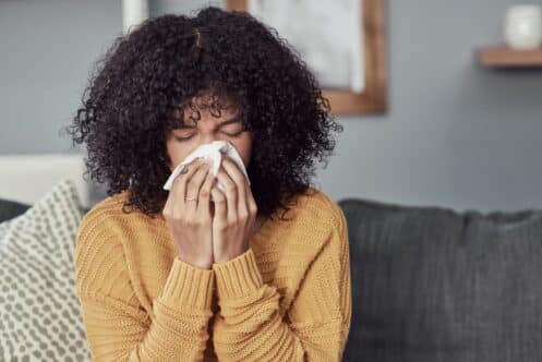 A person with curly hair wearing a yellow sweater sits on a sofa, holding a tissue to their nose, possibly reacting to poor winter indoor air quality. The background features a patterned pillow and a softly blurred interior. | High 5 Plumbing, Heating & Cooling