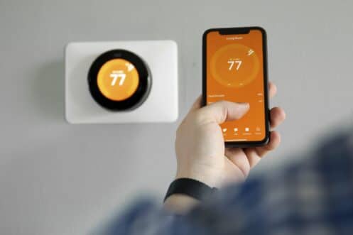 A person holds a smartphone displaying a thermostat app set to 77 degrees, while a smart thermostat on the wall behind them also shows 77 degrees—perfect for year-round comfort. | High 5 Plumbing, Heating & Cooling