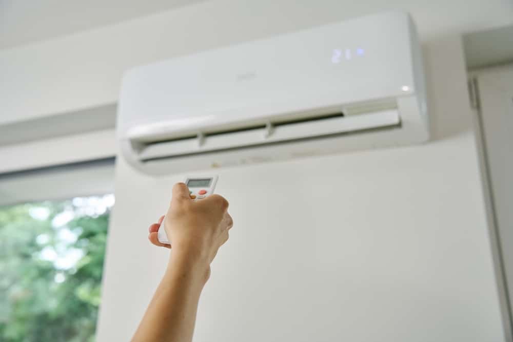 A person points a remote control at a wall-mounted air conditioner, showcasing ductless cooling set to 21 degrees Celsius. A window with greenery outside is partially visible on the left side of the image. | High 5 Plumbing, Heating & Cooling